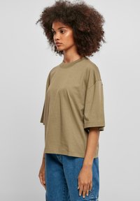 Olive zelené oversized tričko vyrobené z měkké bavlny, s krátkými rukávy a kulatým výstřihem, kombinované s modrými džíny.