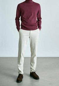 Bordeauxfarbener Rollkragenpullover, weiße Hose und braune Wildlederschuhe. Glattes Material, taillierte Passform und minimalistischer Stil. Schlichter Hintergrund.