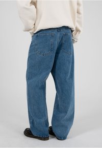 Persoană purtând blugi albaștri largi și o bluză crem, stând cu mâinile în buzunare, pantofi negri vizibili.