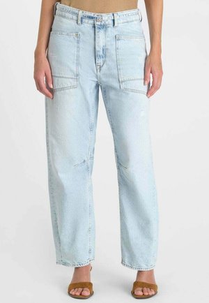 Persoon draagt lichtblauwe jeans met rechte pijpen, grote voorzakken en bruine sandalen met open tenen, staand tegen een effen achtergrond.