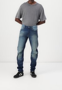 Muž v proschlých modrých džínách, šedém tričku s krátkým rukávem a černých teniskách s bílými pruhy, stojící na bílém pozadí.