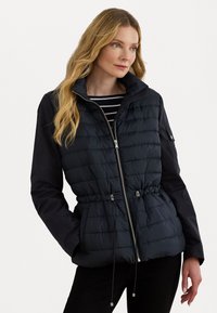 Opblaasjas met een donkerblauw gequilt lichaam, een aansluitende taille, verstelbare koordjes en zwarte lange mouwen. Rits aan de voorkant en zijzakken.
