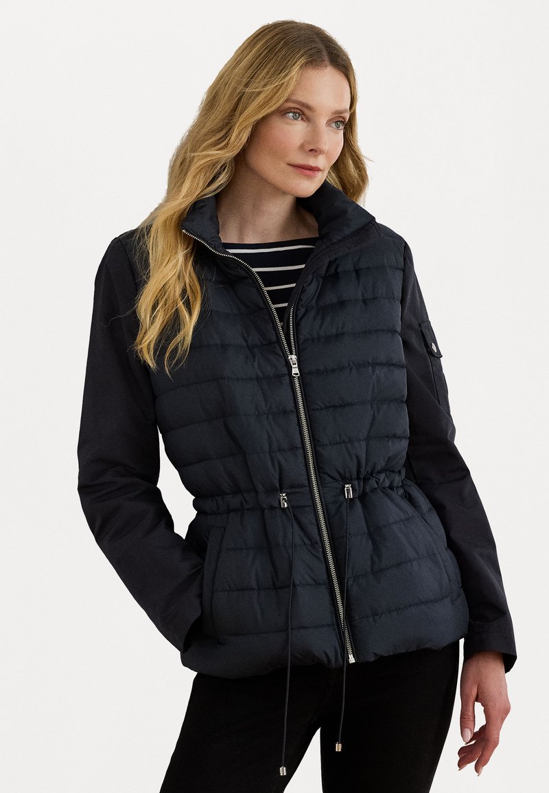Opblaasjas met een donkerblauw gequilt lichaam, een aansluitende taille, verstelbare koordjes en zwarte lange mouwen. Rits aan de voorkant en zijzakken.