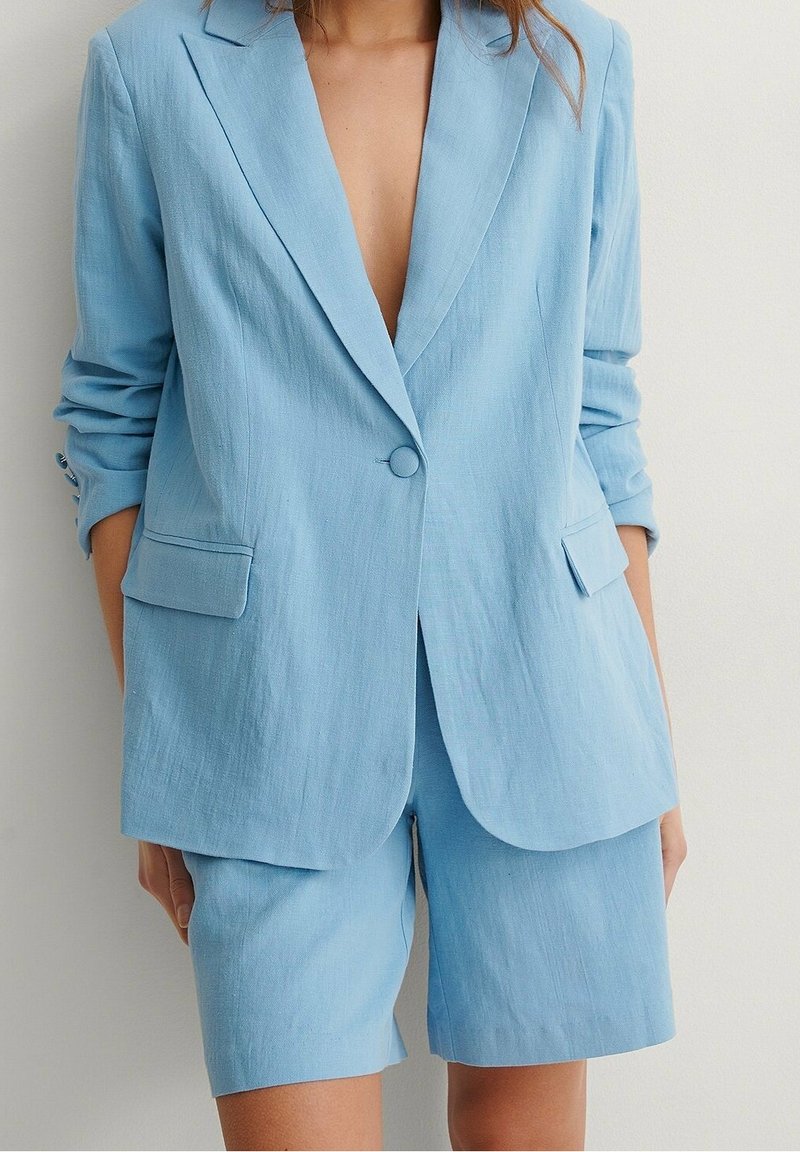 Blazer en lin bleu clair avec des revers structurés, fermeture à un bouton et deux poches avant, associé à un short ajusté assorti.
