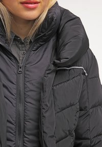 Chaqueta acolchada negra con cuello alto, cierre de cremallera y patrón acolchado. Presenta una tela suave y un pequeño bolsillo en la manga.