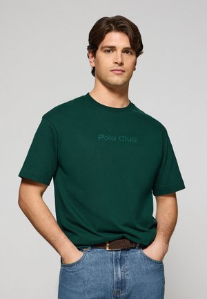 Donkergroene katoenen t-shirt met korte mouwen met "Polo Club" in een lichtere tint, gecombineerd met lichtblauwe denim jeans en een bruine riem.