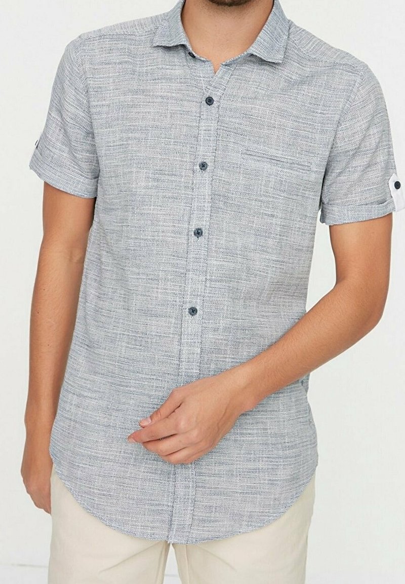 Camisa de manga corta con botones en gris claro y tela texturizada. Tiene un cuello, bolsillo en el pecho y botones oscuros.