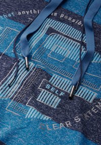 Blaues Hoodie mit Grafikdesign, das verschiedene Texturen, kontrastierende blaue und dunkle Farbtöne sowie Metallspitzen an den Kordeln aufweist.