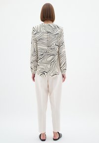 Langærmet beige bluse med et sort bølget mønster, parret med cremefarvede tapered bukser og sorte slip-on sko, set bagfra.