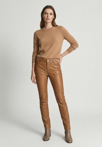 Vrouw die een beige trui, bruine leren broek en taupekleurige enkellaarsjes draagt, staand met één hand op haar heup tegen een effen achtergrond.