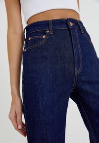 Jeans de tiro alto azul oscuro con costuras de cobre, cinco bolsillos y cierre de botón. Textura de denim suave con un diseño ajustado.