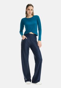 Langärmeliges türkisfarbenes Oberteil mit kurzem Vorderteil, kombiniert mit marineblauen weiten Hosen, die eine glatte Textur und Seitentaschen haben. Weiße Schuhe.