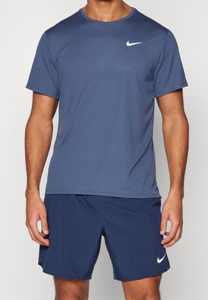Homme portant un t-shirt de sport Nike bleu marine et un short assorti avec des logos Nike blancs, montré du cou jusqu'au milieu de la cuisse, debout sur un fond blanc.