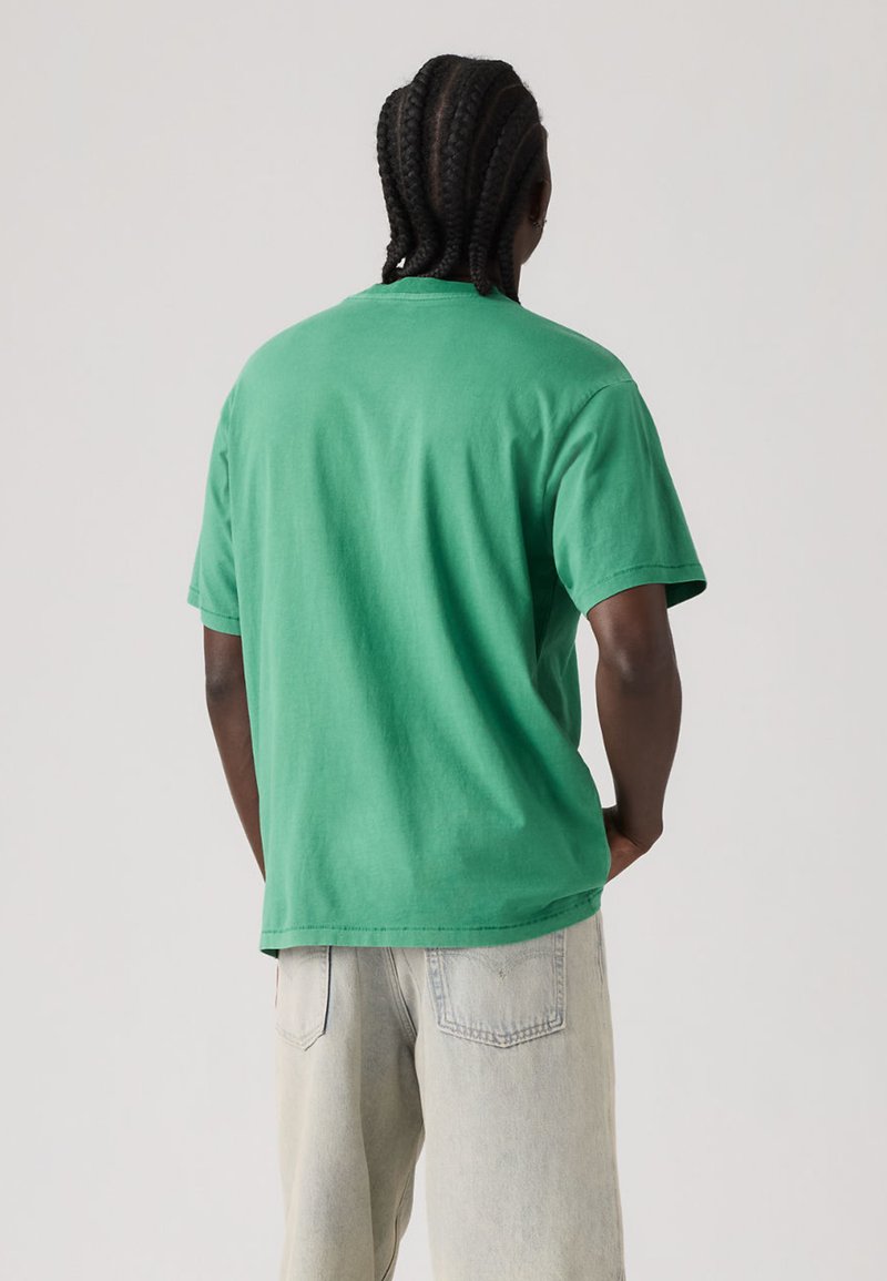 Homme aux cheveux tressés portant un t-shirt vert à manches courtes et un jean de couleur claire, vu de dos sur un fond uni.