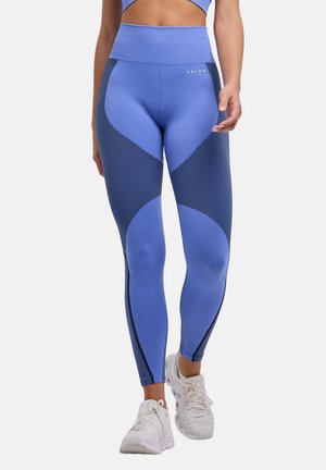 Niebieskie legginsy z ciemnymi wstawami z wzorem na udach i łydkach, zestawione z białymi butami sportowymi na stojącej osobie.