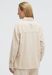 Beige Cordjacke aus Baumwollcord mit lässigem Schnitt, Kragen und einem Etikett auf der Rückseite. Sie verfügt über dezente vertikale Rippungen und Knopfmanschetten.