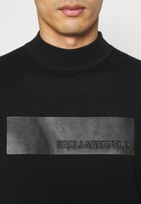 Man med svart ribbad tröja med läderpatch präglad med "KARL LAGERFELD" på bröstet, närbild på hals och mun.