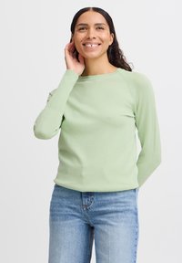 Maglione verde chiaro a maniche lunghe con scollatura tonda e polsini a coste, abbinato a jeans blu chiaro. Texture liscia, design aderente.