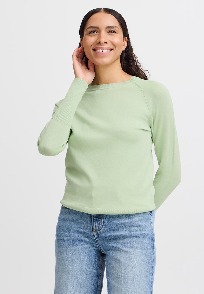 Maglione verde chiaro a maniche lunghe con scollatura tonda e polsini a coste, abbinato a jeans blu chiaro. Texture liscia, design aderente.