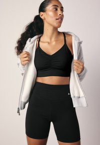Zwarte sportbeha met geplooide details, gecombineerd met hoge zwarte shorts. Het model draagt een grijze rits-geopende hoodie.