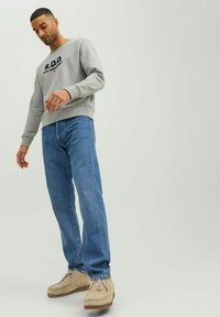 Grauer Sweatshirt mit "R.D.D."-Logo, blaue Jeans und hellbeige Mokassins. Lässiges Outfit an einer stehenden Person.