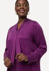 Paarse blouse met lange mouwen, voorzien van een strikdetail bij de hals en een gladde textuur. Heeft een relaxte pasvorm en knopenmanchetten. Geen zichtbare patronen of hardware.