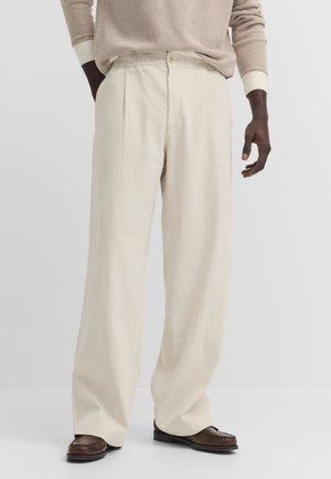 Mann trägt hellbeige weite Hosen, braune Loafers und einen strukturierten beigen Langarmpullover, steht vor einem schlichten Hintergrund.