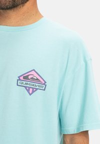 Light blue cotton t-shirt featuring a graphic logo in pink and purple on the left chest area. Short sleeves, relaxed fit, and crew neck.
