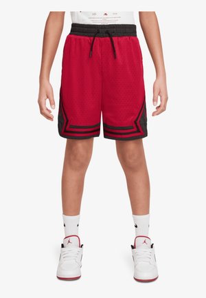 Enfant portant un short de basket rouge et noir, des chaussettes blanches avec des logos noirs et des baskets blanches avec des touches rouges et noires, debout.