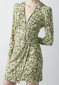 Robe à manches longues avec un motif abstrait vert et marron. Elle présente un décolleté en V profond, des détails froncés à la taille et une silhouette ajustée.