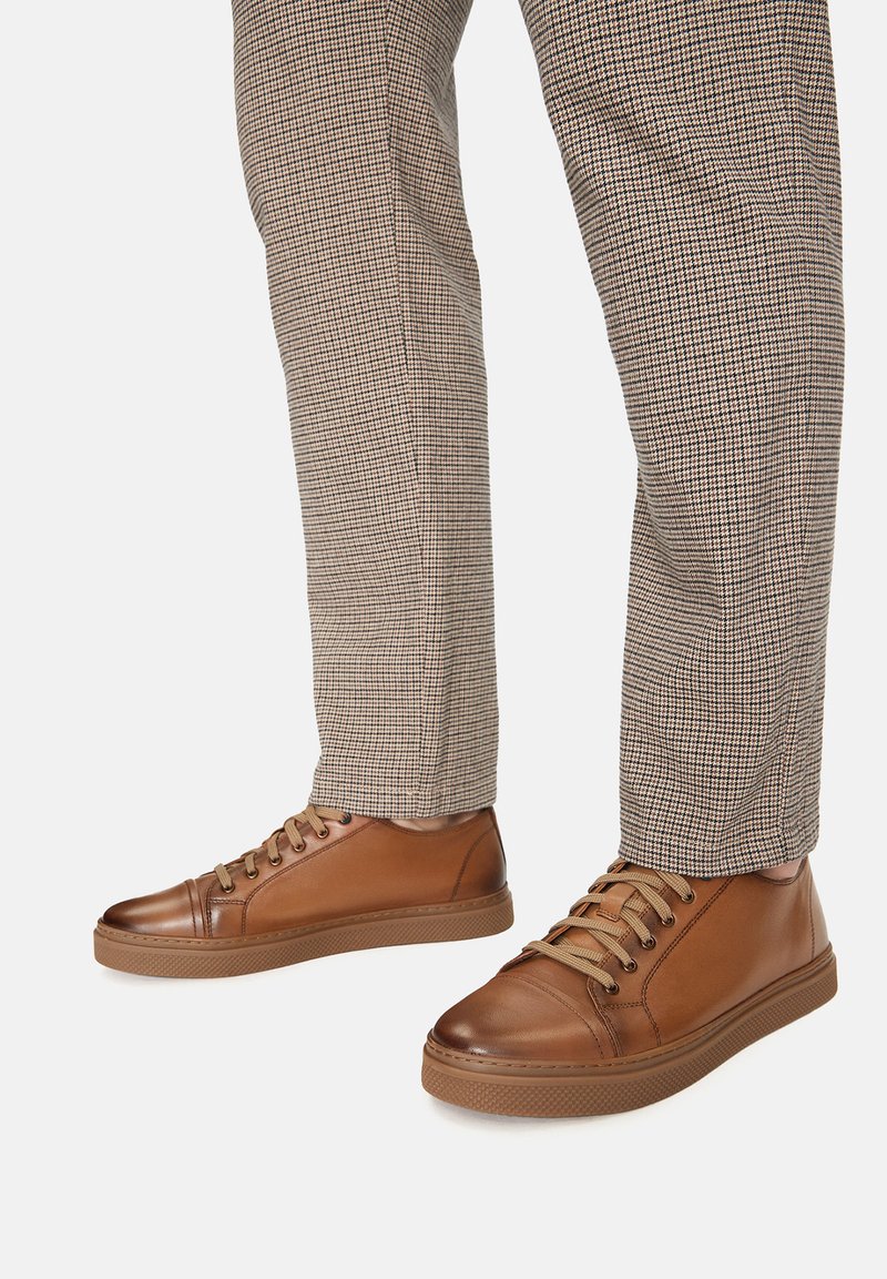 Zapatillas de cuero marrón con una textura suave, que presentan seis ojales, cordones en contraste y una suela de goma, combinadas con pantalones de cuadros.