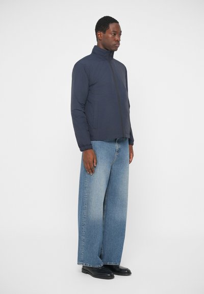 Ναυτική μπλε τζάκετ με φερμουάρ και ψηλό γιακά, υφασμάτινη υφή, φορεμένο με ανοιχτό μπλε φαρδύ παντελόνι και μαύρα παπούτσια. Προφίλ όρθιας στάσης.