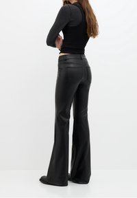 Pantalons synthétiques noirs évasés avec une texture lisse, poches arrière et taille haute. Associés à un haut noir ajusté, vus de dos.