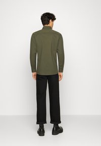 Oliwkowa zielona koszula z długim rękawem i ostrym kołnierzykiem. Tylna część ma przeszycia na ramionach i prosty, schludny wygląd. Do tego czarne, podwinięte spodnie i buty.