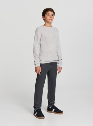 Ragazzo adolescente in piedi, indossa un maglione lavorato a maglia di colore grigio chiaro, pantaloni grigio scuro e scarpe da ginnastica nere con strisce bianche su uno sfondo neutro.