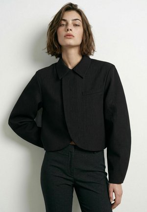 Jeune femme aux cheveux courts et bruns, portant une veste noire courte et ajustée ainsi qu’un pantalon foncé, debout sur un fond blanc uni.