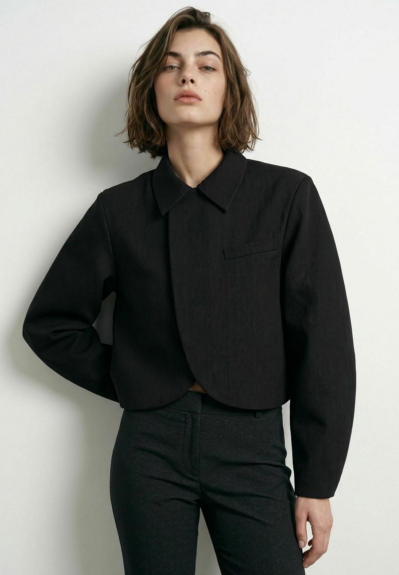 Jeune femme aux cheveux courts et bruns, portant une veste noire courte et ajustée ainsi qu’un pantalon foncé, debout sur un fond blanc uni.
