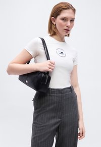 Νεαρή γυναίκα που φοράει λευκό μπλουζάκι με λογότυπο, γκρι παντελόνι με ρίγες και χρυσά σκουλαρίκια κρίκους, κρατώντας μια μαύρη τσάντα ώμου.