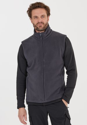 Mørkegrå fleecevest med høj krave, frontlynlås og sidelommer, båret over en sort langærmet skjorte. Glat tekstur, afslappet design.