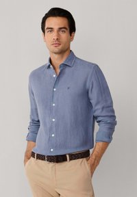 Homme portant une chemise bleue à manches longues avec les poignets retroussés, un pantalon beige et une ceinture tressée foncée, debout les mains dans les poches.
