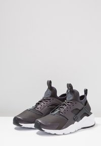 Mustat ja harmaat kengät, joissa on verkkokangas ja synteettiset materiaalit, varustettuna pehmustetulla nilkkareunalla, vetotapeilla, nauhakytkennällä ja valkoisella välikannella.