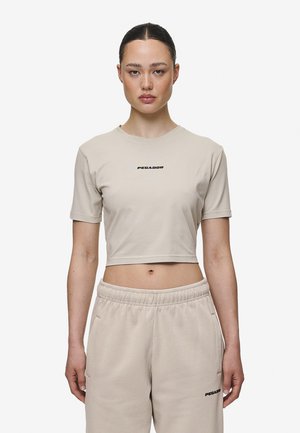 T-shirt curta bege em algodão macio com mangas curtas, apresentando um logo preto "PEGADOR" no peito. Usada com calças de moletom bege a condizer.