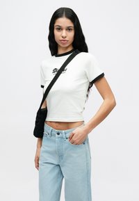 Νεαρή γυναίκα με μακριά μαύρα μαλλιά που φοράει λευκό κοντομάνικο τοπ, ανοιχτό μπλε τζιν και μαύρη τσάντα χιαστί, στέκεται μπροστά σε απλό φόντο.