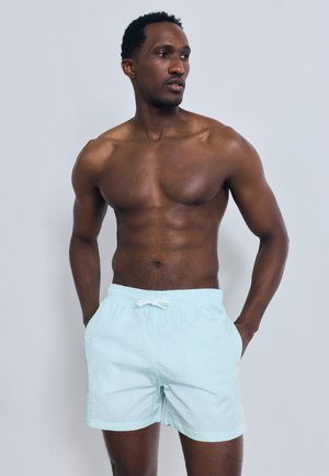 Μυώδης άνδρας με σκούρο δέρμα που φοράει ανοιχτό μπλε μαγιό, στέκεται με τα χέρια στις τσέπες, κοιτάζοντας προς τα αριστερά του με φόντο ένα απλό σκηνικό.