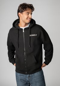 Black zip-up hoodie made of soft fabric, featuring a front pocket, drawstrings, and white text with "LOW IMPACT" on the left chest.