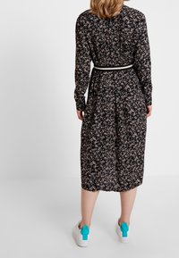 Czarna sukienka w kwiaty z długimi rękawami, doskonale podkreślająca talię, o długości do połowy łydki. Noszona z białymi sneakersami z turkusowymi akcentami.