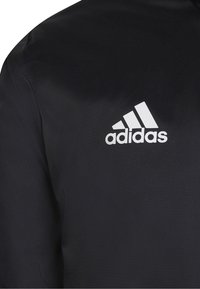 Schwarze Adidas-Jacke aus strukturiertem Stoff; mit einem auffälligen weißen Logo-Design auf der oberen linken Seite.