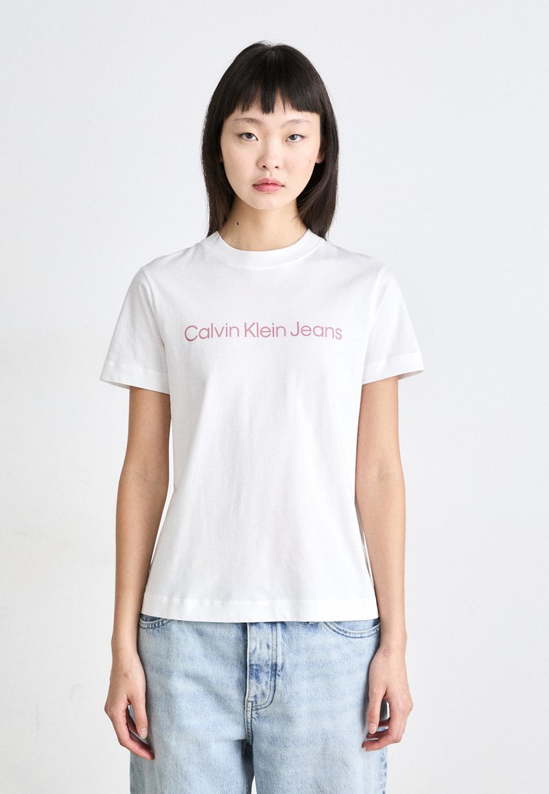 Biela bavlnená tričko s okrúhlym výstrihom a krátkymi rukávmi, s nápisom "Calvin Klein Jeans" v svetloružovej farbe naprieč prsiami.