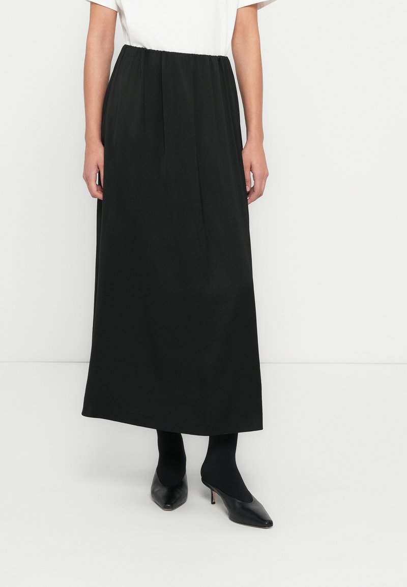 Jupe longue noire avec une ceinture élastique, tissu uni, design fluide, présentée avec une chemise blanche et des chaussures noires.