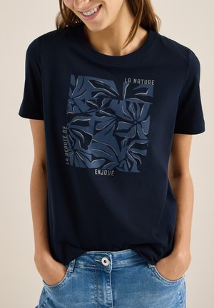 Femme souriante portant un t-shirt bleu marine avec un imprimé abstrait de feuilles bleu foncé et un texte en français, associée à un jean bleu clair, les mains dans les poches.